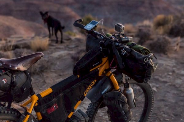 A loaded bike stands on a rocky terrain with a dog nearby, overlooking a vast canyon at sunset with colorful skies in the background.