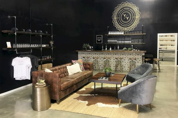 A stylish lounge area features a brown leather sofa, two grey chairs, a coffee table, and a decorative bar against a black wall with shelves.