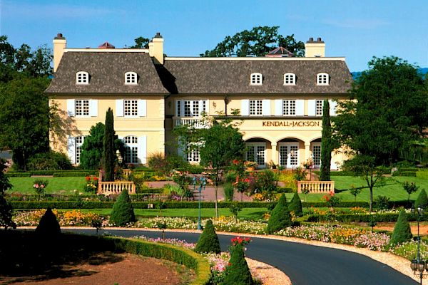 The image shows a grand house with a neatly manicured garden and a curved driveway leading up to the entrance, surrounded by lush greenery.