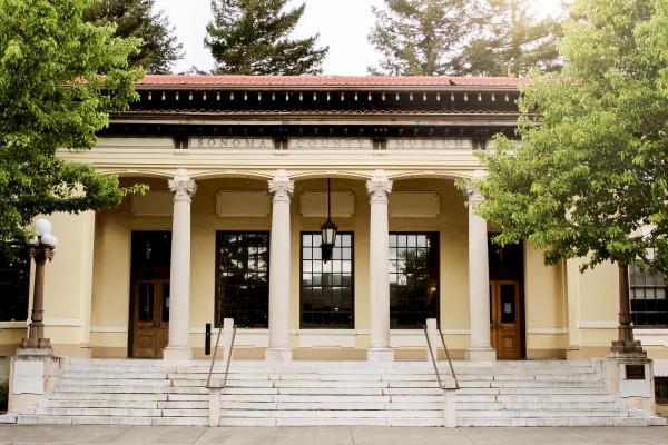 The image shows a building with four large columns, steps leading up to the entrance, flanked by two doors, with trees in the background.