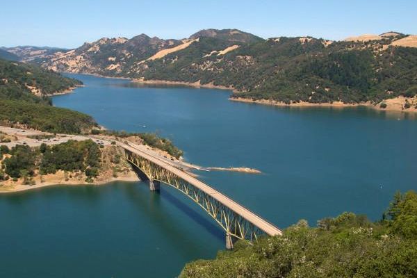 The image depicts a scenic view of a bridge spanning across a serene lake, surrounded by lush green hills and mountains under a clear blue sky.