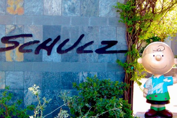 A statue of a smiling cartoon character stands next to a wall with "SCHULZ" written on it in large letters, surrounded by greenery and plants.