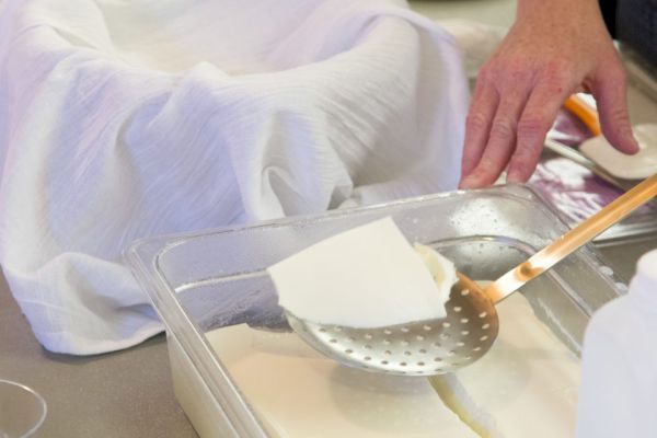 A person is preparing tofu, using a slotted spoon to lift a piece from a container, with a cloth-lined bowl in the background.