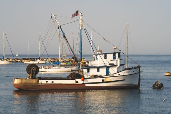 A fishing boat with an American flag docked in calm waters, alongside other smaller boats in the background, under a clear sky.