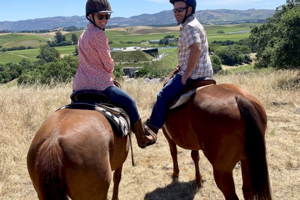 Two people wearing helmets and casual clothing are horseback riding in an open field with scenic hills and greenery in the background.