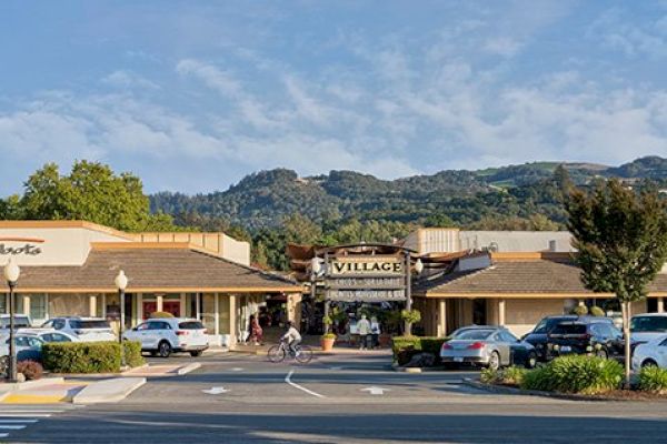 A shopping center called "VILLAGE" with parked cars, mountains in the background, and a few people walking around the area, during the day.