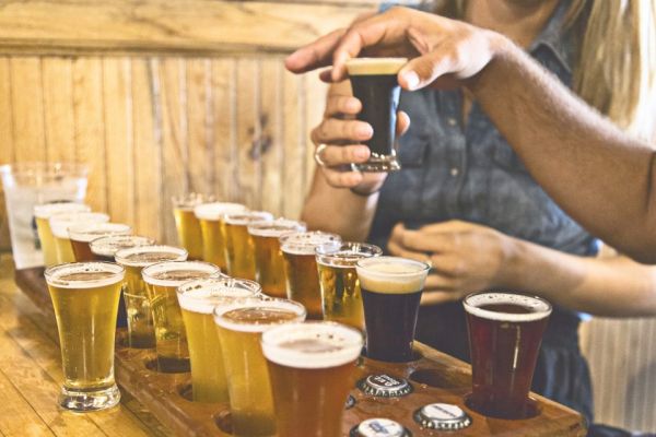 Several glasses of different types of beer on a wooden tray, with two people sampling them. Some bottle caps are visible on the table.