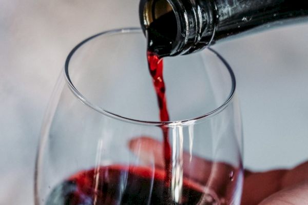 A bottle is pouring red wine into a glass, with a hand holding the glass, positioned against a blurred background.