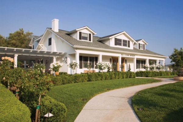 A large white house with dormer windows, a wraparound porch, and well-maintained landscaping, including hedges and a curved pathway ending the sentence.