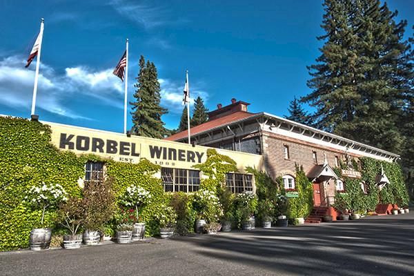 The image shows the exterior of the Korbel Winery, a building with green ivy, flags, potted plants, and a blue sky with trees in the background.