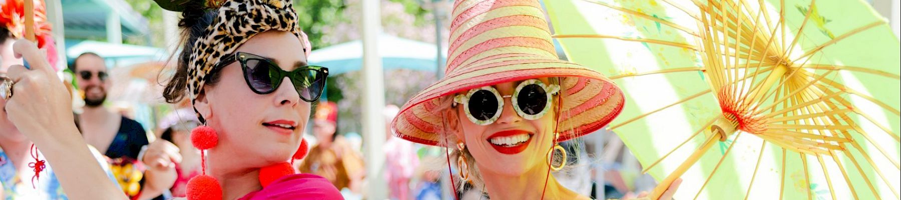 Two women dressed in colorful, floral attire with sunglasses and hats, holding umbrellas, enjoying a sunny day outdoors.