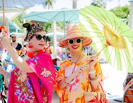 Two women dressed in colorful, floral attire with sunglasses and hats, holding umbrellas, enjoying a sunny day outdoors.