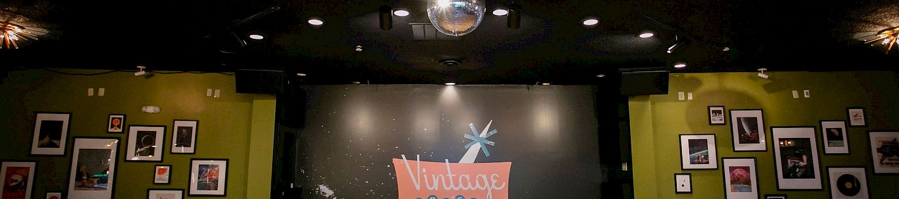 The image depicts a small stage in a vintage-themed venue with framed photos on green walls and a disco ball hanging from the ceiling.