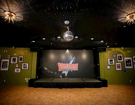 The image depicts a small stage in a vintage-themed venue with framed photos on green walls and a disco ball hanging from the ceiling.