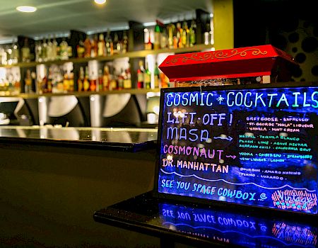 A neon sign at a bar advertises "Cosmic Cocktails" including drinks like Liftoff, Masa, Cosmonaut, and Dr. Manhattan. See you space cowboy.