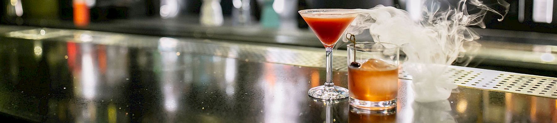 A bar counter with assorted alcoholic beverages in the background, featuring two cocktails, one in a martini glass and one with smoke.