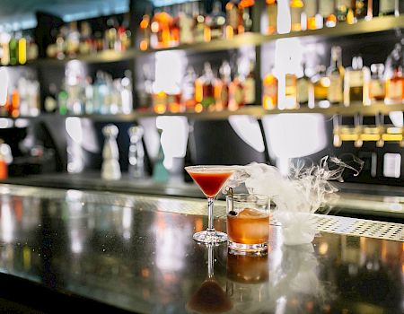 A bar counter with assorted alcoholic beverages in the background, featuring two cocktails, one in a martini glass and one with smoke.