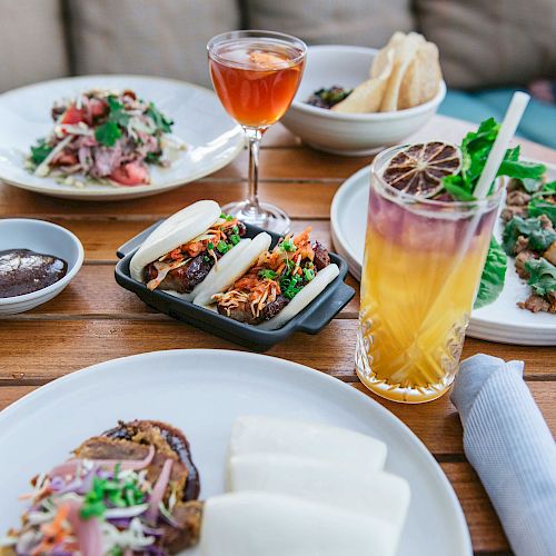 The image shows a wooden table with various dishes including tacos, a colorful cocktail, a salad, and other plates, with a napkin on the side.