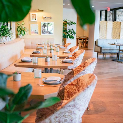 A stylish, well-lit restaurant interior with neatly set tables, cushioned chairs, plants, and large windows letting in natural light.