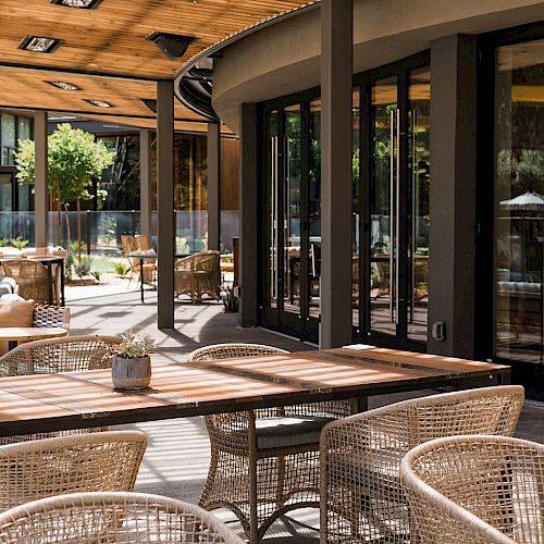 An outdoor seating area with wooden tables, wicker chairs, and cushioned sofas, adjacent to a building with large glass windows and surrounded by greenery.