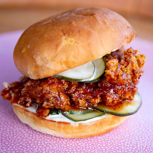 The image displays a fried chicken sandwich with pickles and sauce, served on a bun, placed on a pink plate with a speckled design, ready to be eaten.