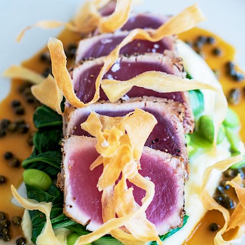 The image shows a gourmet dish of seared tuna slices on a bed of greens, garnished with crispy onion strings, and surrounded by a sauce with capers.