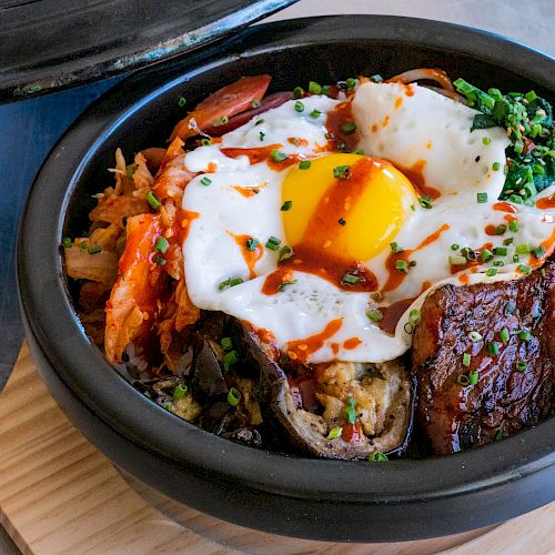 A bowl of bibimbap, a Korean dish with rice, vegetables, kimchi, beef, and a fried egg topped with chili sauce and chopped chives.