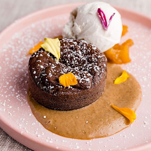 A chocolate lava cake garnished with powdered sugar and floral petals, served with a scoop of vanilla ice cream and a caramel sauce on a pink plate.