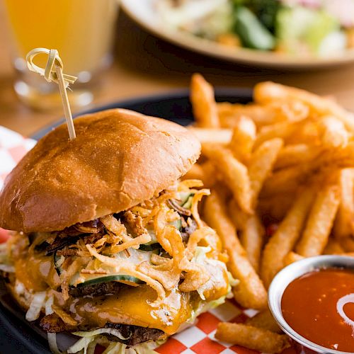 A delicious-looking burger with fries and a side of ketchup; a beverage and salad are blurred in the background, creating a tasty meal scene.