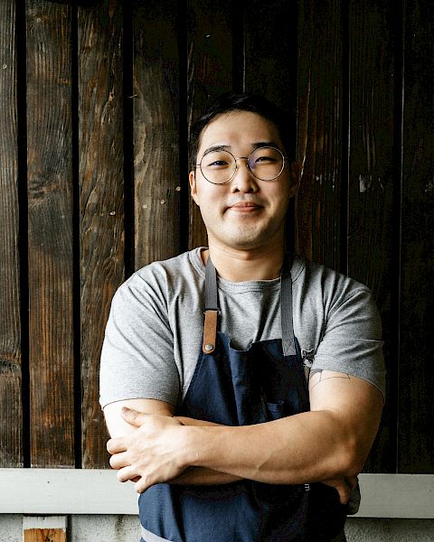 A person wearing glasses and an apron stands confidently with crossed arms in front of a wooden backdrop and a partial brick wall.