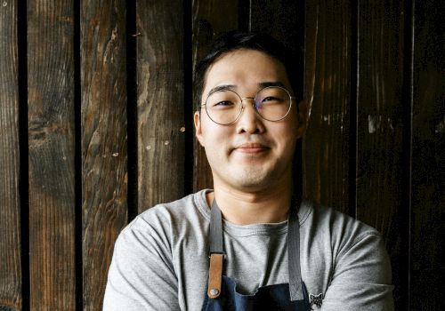 A person wearing glasses and an apron stands confidently with crossed arms in front of a wooden backdrop and a partial brick wall.