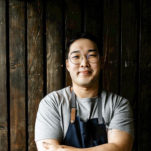 A person wearing glasses and an apron stands confidently with crossed arms in front of a wooden backdrop and a partial brick wall.