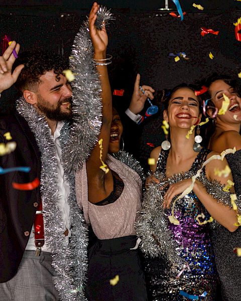 A group of people celebrate, wearing festive attire and tinsel, while confetti falls around them in a joyous setting.