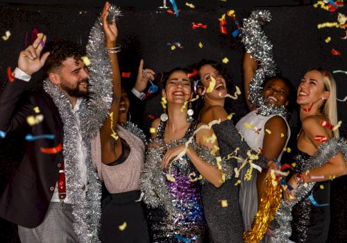 A group of people celebrate, wearing festive attire and tinsel, while confetti falls around them in a joyous setting.