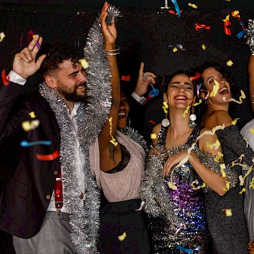 A group of people celebrate, wearing festive attire and tinsel, while confetti falls around them in a joyous setting.