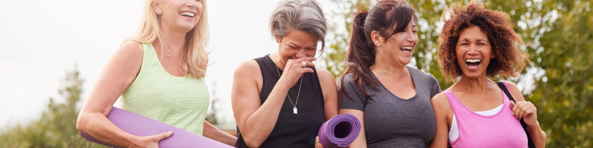 Four women are walking outdoors, laughing and chatting, each holding a yoga mat, dressed in workout clothing with a natural background.