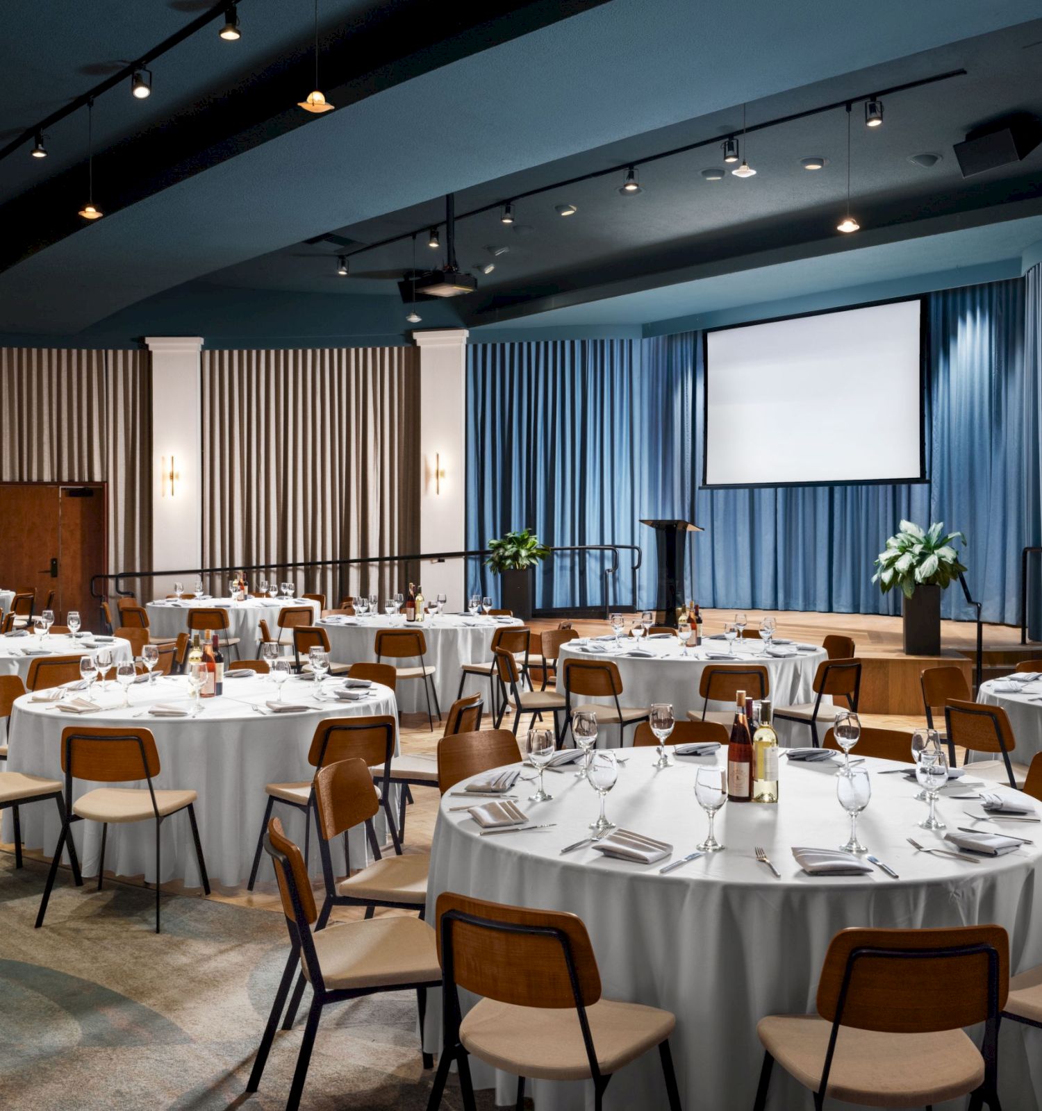 The image shows a banquet hall with round tables, white tablecloths, chairs, and table settings, set up for an event. A stage with a screen is at the front.