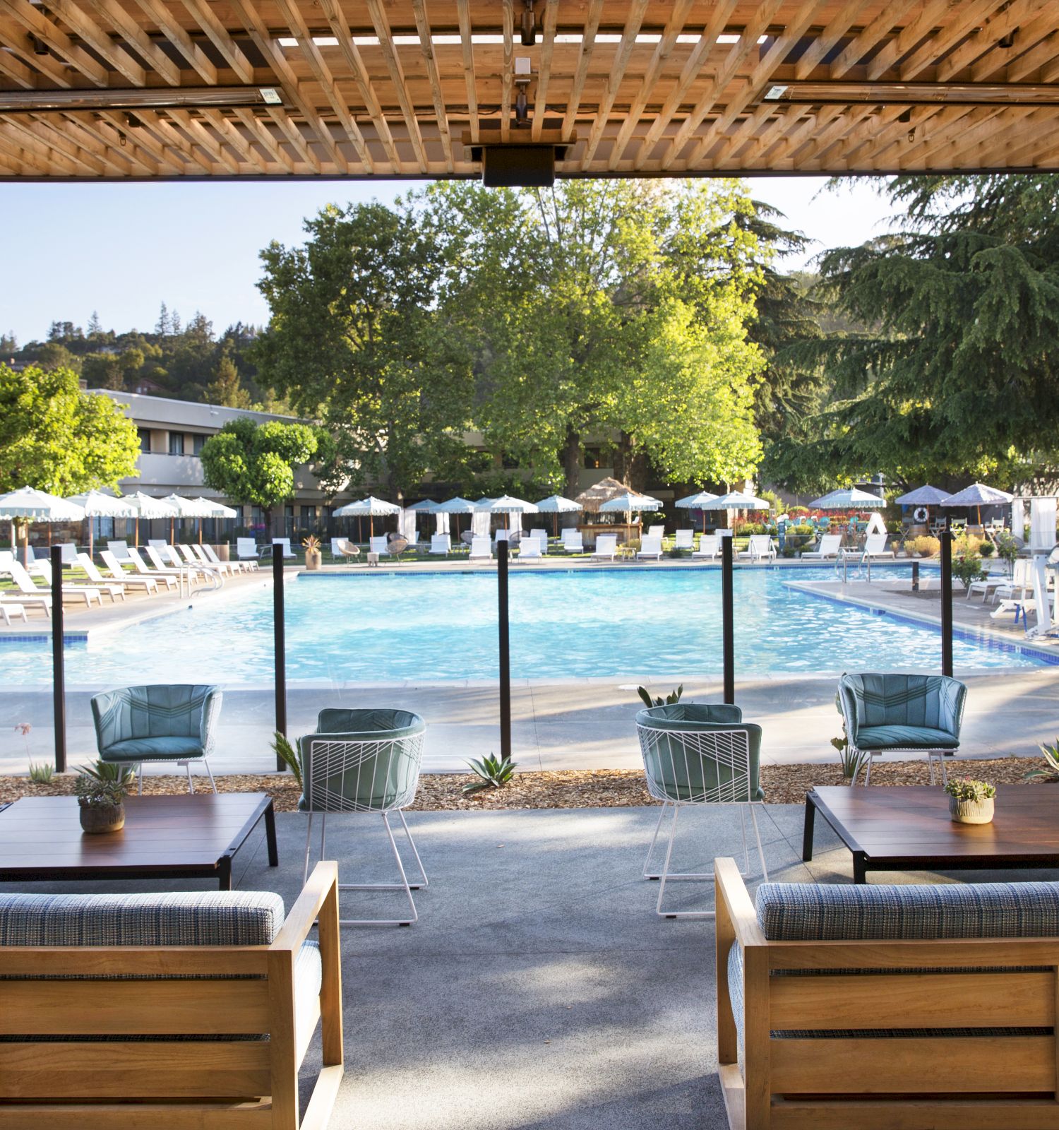 An outdoor seating area with sofas and tables overlooks a swimming pool surrounded by lounge chairs and umbrellas, with trees and greenery in the background.