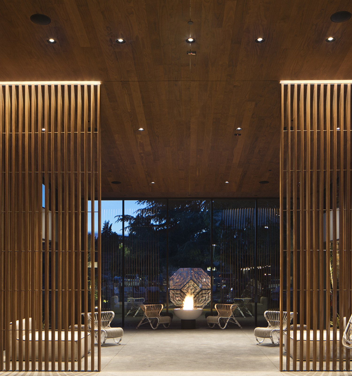 A modern outdoor seating area with wooden slat structures, hanging chairs, and a lit pathway leading to a garden in the evening.