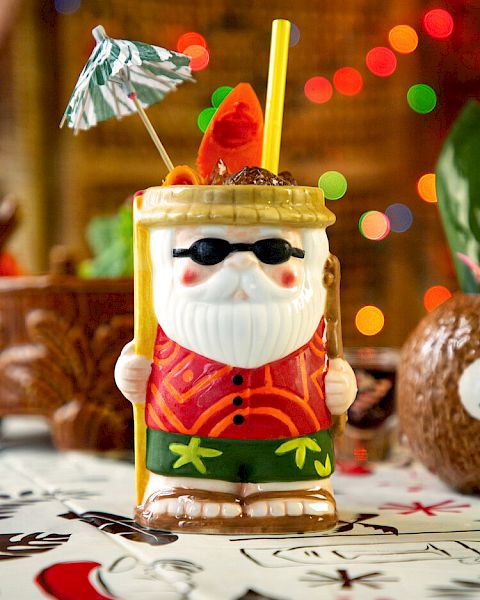 A festive Santa-themed tiki drink is decorated with a small umbrella and straw, with colorful lights in the background, creating a tropical holiday vibe.