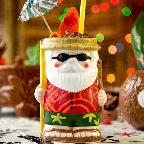 A festive Santa-themed tiki drink is decorated with a small umbrella and straw, with colorful lights in the background, creating a tropical holiday vibe.