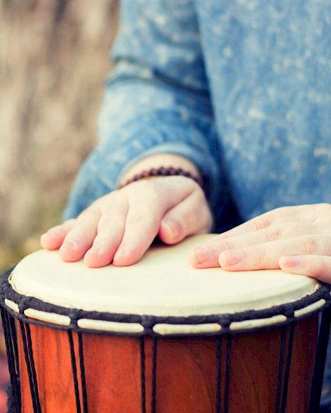 The image shows hands tapping on a djembe drum, with the person wearing a blue shirt and beaded bracelets.