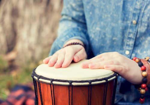 The image shows hands tapping on a djembe drum, with the person wearing a blue shirt and beaded bracelets.