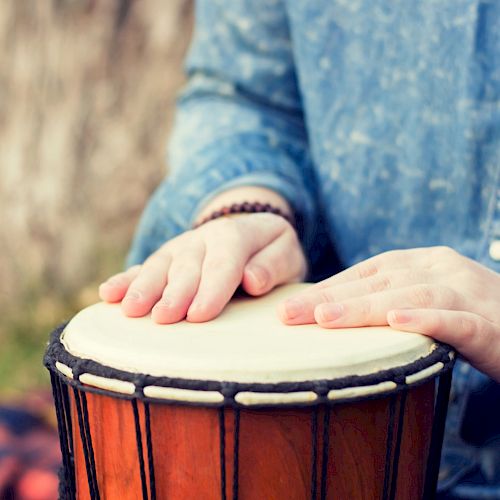 The image shows hands tapping on a djembe drum, with the person wearing a blue shirt and beaded bracelets.