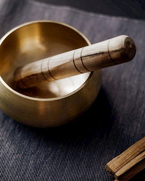 The image shows a Tibetan singing bowl with a wooden striker resting on a fabric surface, alongside a bundle of incense sticks tied with a green string.