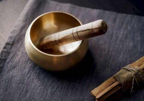 The image shows a Tibetan singing bowl with a wooden striker resting on a fabric surface, alongside a bundle of incense sticks tied with a green string.