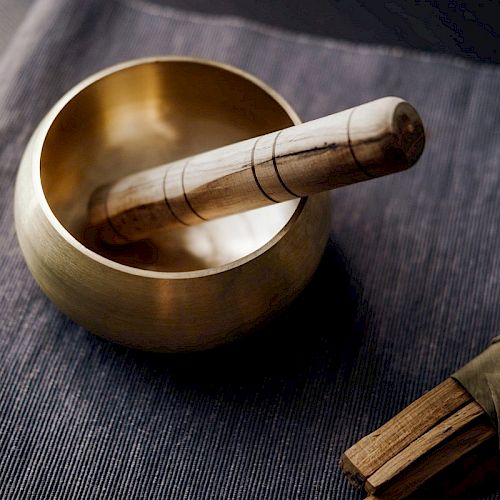 The image shows a Tibetan singing bowl with a wooden striker resting on a fabric surface, alongside a bundle of incense sticks tied with a green string.