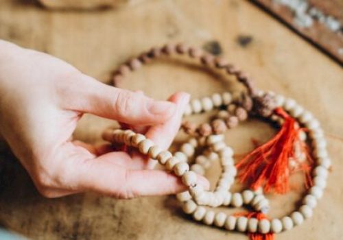 A hand is holding wooden prayer beads with a red tassel, laid out on a wooden surface. Nearby is an incense holder.