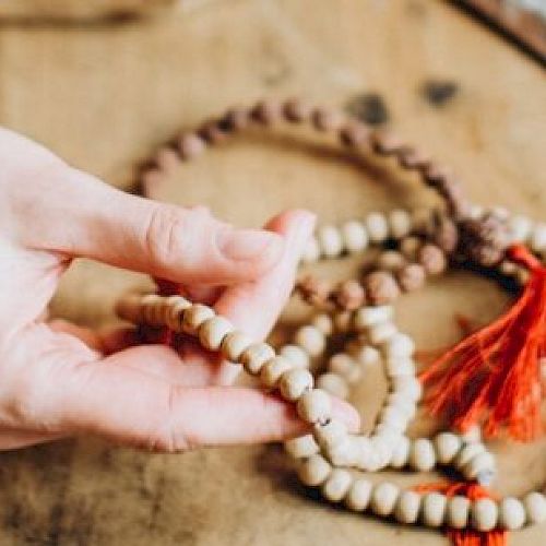 A hand is holding wooden prayer beads with a red tassel, laid out on a wooden surface. Nearby is an incense holder.