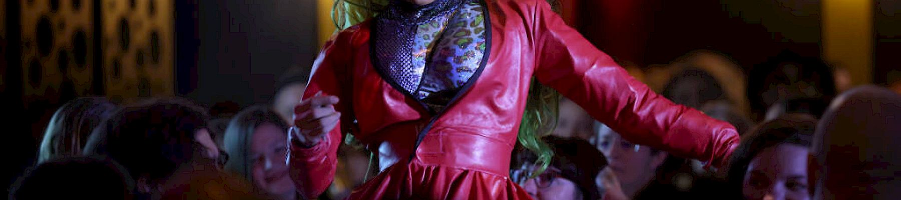 A performer with vibrant yellow hair and a red outfit entertains a crowd in what appears to be a lively indoor venue.